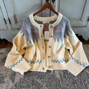 May include: Cream-coloured cardigan sweater with a round neckline and button-up front. The sweater features a geometric pattern in shades of blue and pink across the shoulders and sleeves. The sleeves and bottom hem have a zig-zag pattern.
