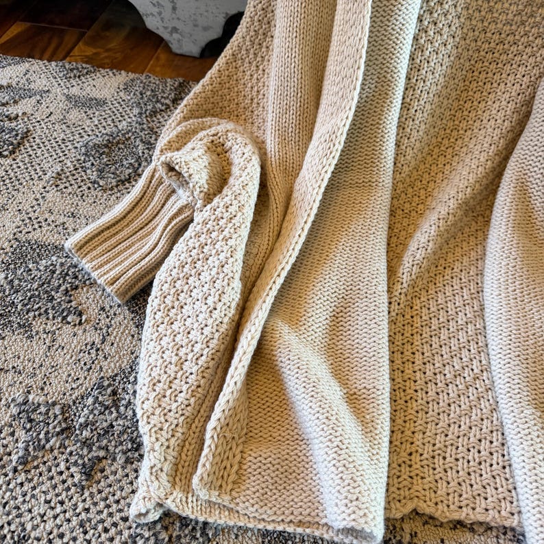 May include: A beige knitted cardigan with a textured pattern. The open-front sweater features long sleeves and a relaxed fit. The soft, neutral color and chunky knit design create a cozy and comfortable aesthetic.