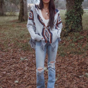 May include: A woman wearing a brown cowboy hat, a gray patterned hooded sweater with fringe, a white top, and light blue ripped jeans. She is standing outdoors in a wooded area with fallen leaves on the ground.
