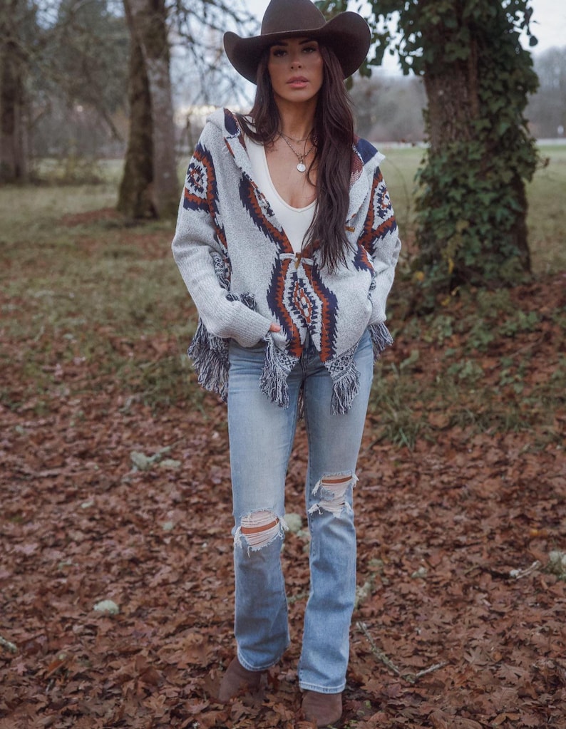 May include: A woman wearing a brown cowboy hat, a gray patterned hooded sweater with fringe, a white top, and light blue ripped jeans. She is standing outdoors in a wooded area with fallen leaves on the ground.