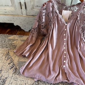 May include: A mauve blouse with long sleeves and a button-up front. The sleeves are made of delicate lace with floral patterns. The blouse has a relaxed fit and a flowy silhouette.