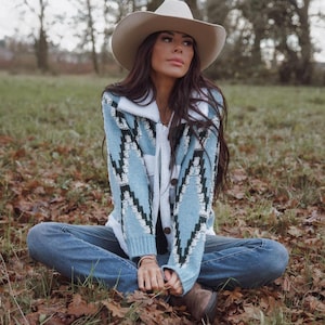 May include: A light blue cardigan with a geometric black and white pattern. The cardigan has a white collar and is buttoned up. The person is wearing a beige cowboy hat and blue jeans while sitting on the ground.