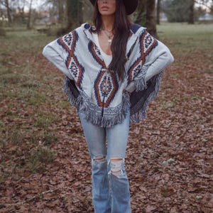 May include: A gray poncho with a geometric pattern in shades of brown and blue, paired with light blue ripped jeans and a brown cowboy hat. The poncho has a fringed hemline. The outfit is worn outdoors.
