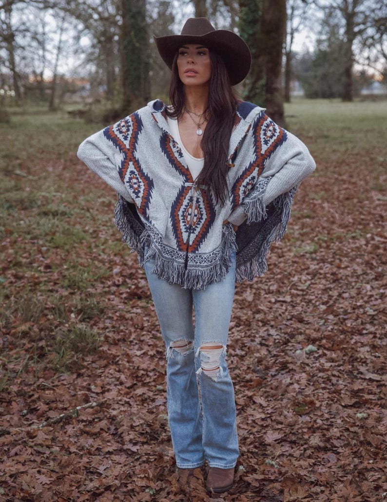 May include: A gray poncho with a geometric pattern in shades of brown and blue, paired with light blue ripped jeans and a brown cowboy hat. The poncho has a fringed hemline. The outfit is worn outdoors.