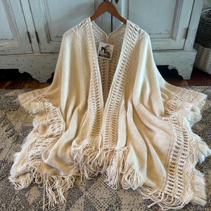 Peut inclure: Châle en tricot crème avec des franges. Le châle présente une conception ouverte sur le devant avec des motifs complexes sur les bords et une frange délicate. Le châle est présenté sur un cintre en bois.