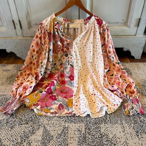 Könnte beinhalten: Eine langärmelige Bluse mit Patchwork-Design, bestehend aus verschiedenen Blumen- und geometrischen Mustern in Rosa, Gelb, Blau und Weiß. Das Kleidungsstück hat einen gerafften Halsausschnitt, eine V-förmige Frontöffnung und elastische Ärmelbündchen.