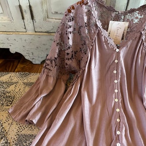 May include: A mauve button-up blouse with long sleeves and a lace overlay on the shoulders. The lace is a floral pattern. The blouse has a "Love, Faith, & Grace" tag attached.
