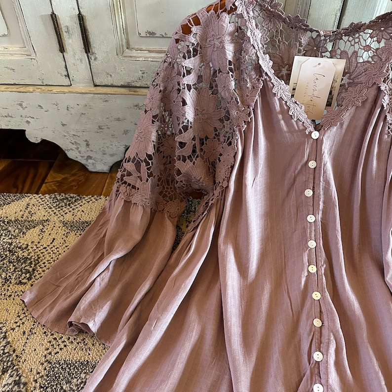 May include: A mauve button-up blouse with long sleeves and a lace overlay on the shoulders. The lace is a floral pattern. The blouse has a "Love, Faith, & Grace" tag attached.