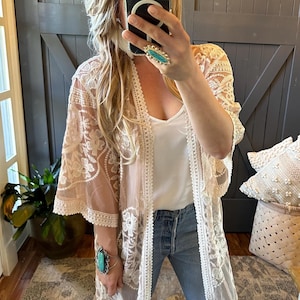 Puede incluir: Kimono de encaje color rubor con patrones florales y ribete blanco. El diseño abierto presenta mangas anchas. El kimono se combina con una camiseta blanca y vaqueros azules. Estética delicada y femenina.