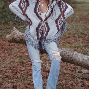 May include: A gray poncho with a geometric pattern in brown and navy blue, featuring fringe trim. The poncho is worn with light blue ripped jeans and a brown cowboy hat. The outfit is completed with brown boots.