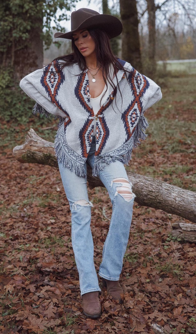May include: A gray poncho with a geometric pattern in brown and navy blue, featuring fringe trim. The poncho is worn with light blue ripped jeans and a brown cowboy hat. The outfit is completed with brown boots.