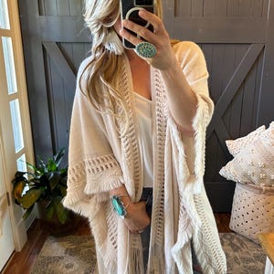 May include: A cream-coloured crochet cardigan with fringe detailing. The cardigan is open at the front and has a loose fit. The model is wearing the cardigan over a white vest top and blue jeans.