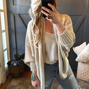 May include: A beige open-front knit cardigan with a textured weave and ribbed cuffs, worn over a white sleeveless top and light blue distressed denim jeans. The outfit is accessorized with a large stone ring and a turquoise cuff bracelet.
