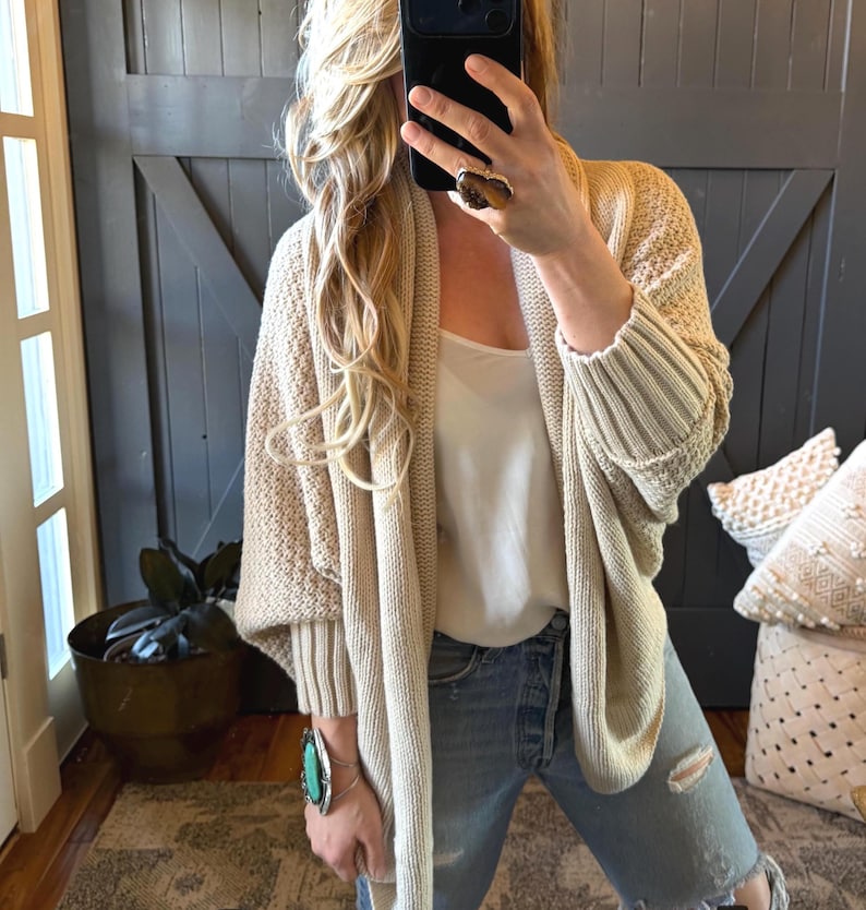May include: A beige open-front knit cardigan with a textured weave and ribbed cuffs, worn over a white sleeveless top and light blue distressed denim jeans. The outfit is accessorized with a large stone ring and a turquoise cuff bracelet.