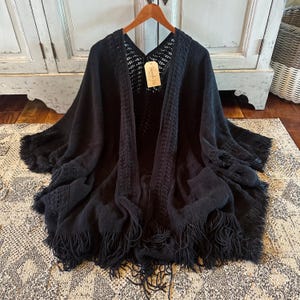 May include: A black knitted poncho with a V-neck design and fringe detailing. The poncho is displayed on a wooden hanger. The open weave design is visible on the upper chest area. A small tag is attached to the poncho.