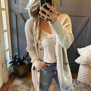 May include: A long, beige cardigan with a ribbed collar and cuffs. The cardigan has two front pockets and is buttoned up. The person is wearing a white camisole and blue jeans.