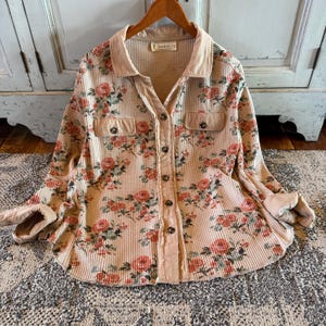 May include: A beige waffle-knit shirt with a floral rose pattern in shades of red, pink, and green. The shirt features a collared neckline, two chest pockets, and button-down closure. The cuffs are rolled up, revealing a lighter beige fabric.