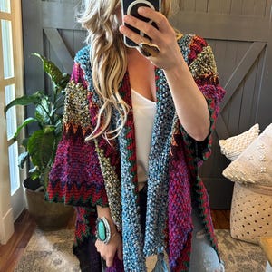 May include: A colorful, multi-patterned knitted poncho with fringe. The poncho is made of a soft, textured yarn and features a variety of geometric designs in shades of blue, green, red, and yellow.