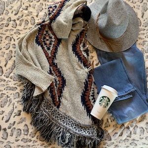 May include: A neutral-toned poncho with a geometric pattern in navy and brown, paired with a gray hat, blue jeans, and a Starbucks coffee cup. The poncho has a fringed hemline and a hood.