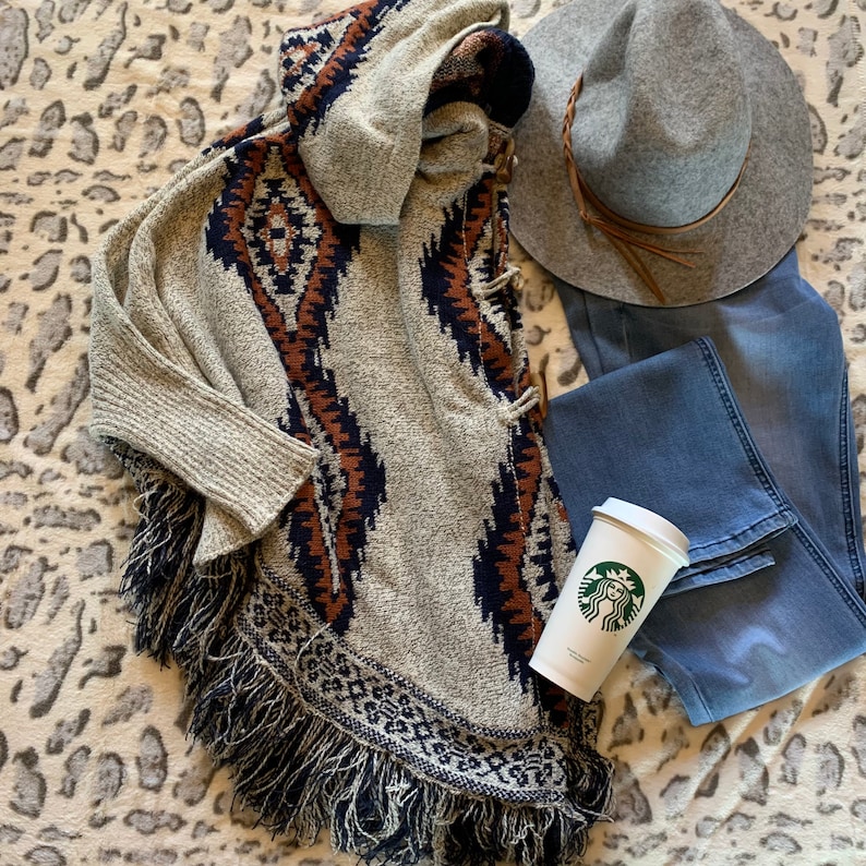May include: A neutral-toned poncho with a geometric pattern in navy and brown, paired with a gray hat, blue jeans, and a Starbucks coffee cup. The poncho has a fringed hemline and a hood.