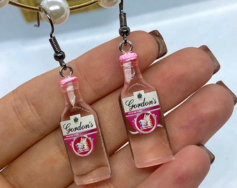 May include: A pair of pink and clear glass bottle earrings with a pink cap and a silver label that reads "Gordon's Premium Pink Distilled Gin" with a pink and white crest.