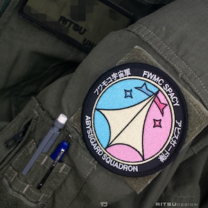 May include: Olive green military-style jacket with a circular patch. The patch features a blue, pink, and beige design with Japanese text and the words "FWMC SPACEY" and "ABYSSGARD SQUADRON". A pen and pencil are in the pocket.