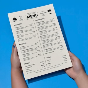 Pode incluir: Um menu de restaurante com opções de pequeno-almoço, prato principal e sobremesa. O menu, segurado por uma pessoa, está contra um fundo azul. Os pratos incluem Ovos Benedict, frango Piccata e bolo de lava de chocolate.