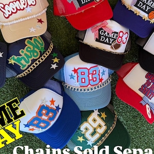 May include: A selection of mesh-backed trucker caps in various colours, with embroidered text and graphics. Designs include team names, numbers, and the phrase "Game Day." The caps have a classic trucker style with a curved peak.