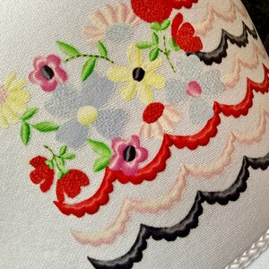 May include: Close-up of a white fabric with a floral design. The design features red, pink, yellow, and blue flowers with green leaves. A scalloped border in red, pink, and black accents the floral arrangement. The fabric has a delicate, textured appearance.