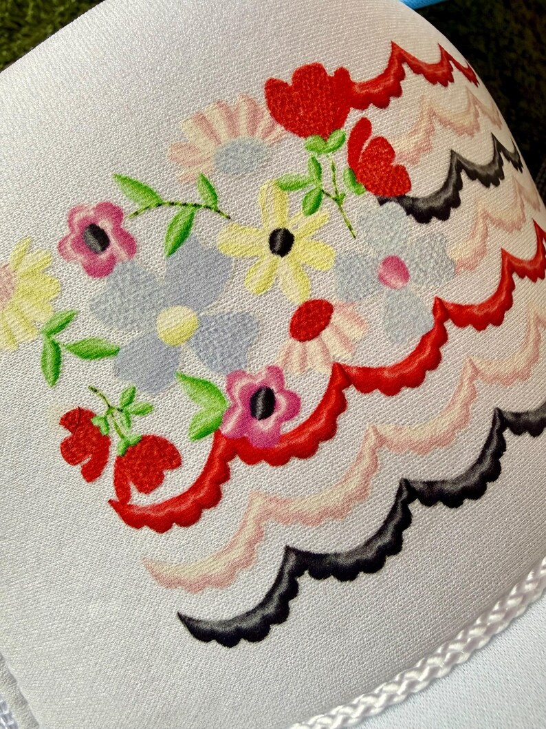 May include: Close-up of a white fabric with a floral design. The design features red, pink, yellow, and blue flowers with green leaves. A scalloped border in red, pink, and black accents the floral arrangement. The fabric has a delicate, textured appearance.
