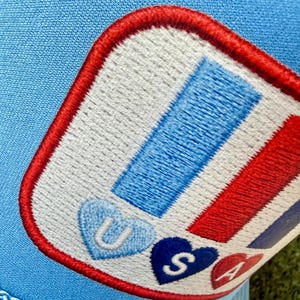 May include: Light blue trucker hat with a patch featuring red, white, and blue stripes and the letters "USA" in heart shapes. The patch is outlined in red and stitched onto the hat.