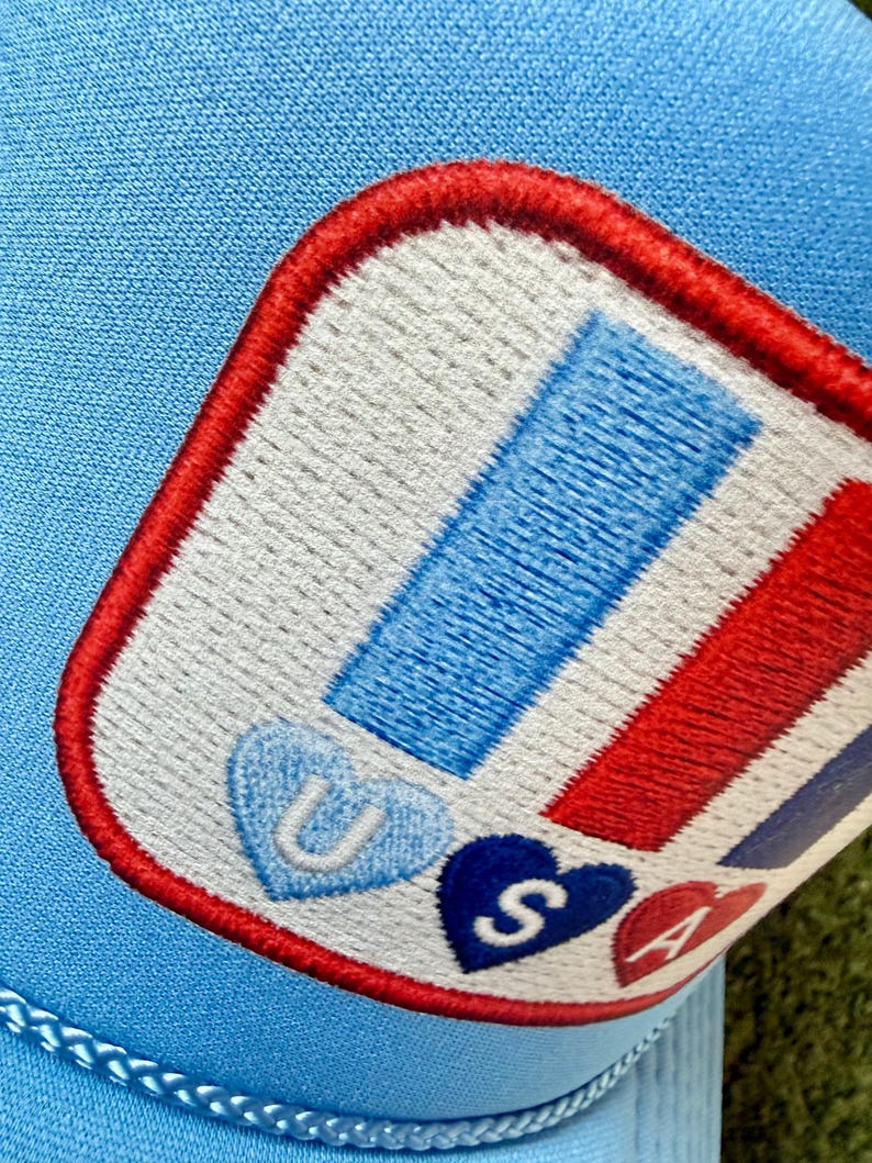 May include: Light blue trucker hat with a patch featuring red, white, and blue stripes and the letters "USA" in heart shapes. The patch is outlined in red and stitched onto the hat.