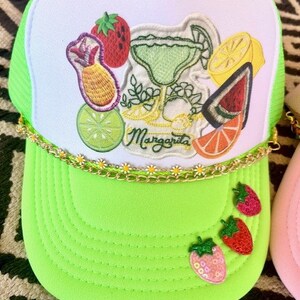 May include: A green and white trucker hat with a margarita and fruit embroidery. The hat has a gold chain trim and the words "Margarita" are embroidered on the front.