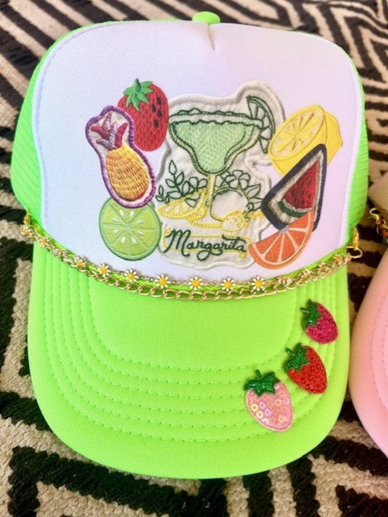 May include: A green and white trucker hat with a margarita and fruit embroidery. The hat has a gold chain trim and the words "Margarita" are embroidered on the front.