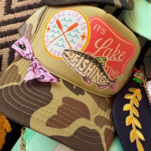 May include: A collection of trucker hats with various designs. One hat features a camouflage pattern, while another has a patch with the text "Lake Life is the Best" and a fish graphic. Other hats have decorative elements.