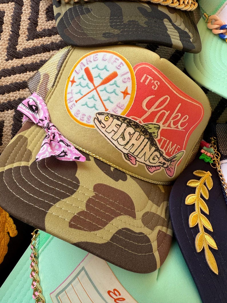 May include: A collection of trucker hats with various designs. One hat features a camouflage pattern, while another has a patch with the text "Lake Life is the Best" and a fish graphic. Other hats have decorative elements.