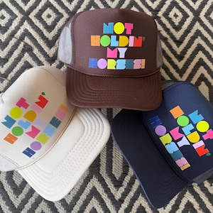 May include: Three trucker hats in brown, white, and navy blue. Each hat features the phrase "NOT HOLDIN' MY HORSES!" in colorful, blocky letters. The hats have a mesh back and a curved brim.