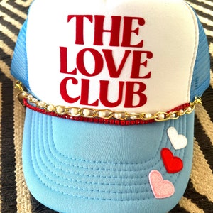 May include: A blue and white trucker hat with a red embroidered logo that says "THE LOVE CLUB". The hat has a gold chain and pearl trim around the brim and three felt hearts on the front.