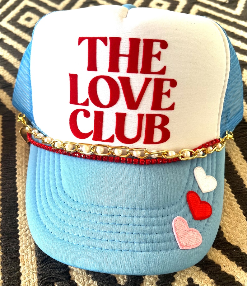May include: A blue and white trucker hat with a red embroidered logo that says "THE LOVE CLUB". The hat has a gold chain and pearl trim around the brim and three felt hearts on the front.