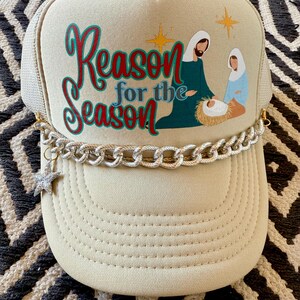 May include: A beige trucker hat with a gold chain trim and a star charm. The front of the hat features a Christmas nativity scene with the text "Reason for the Season".