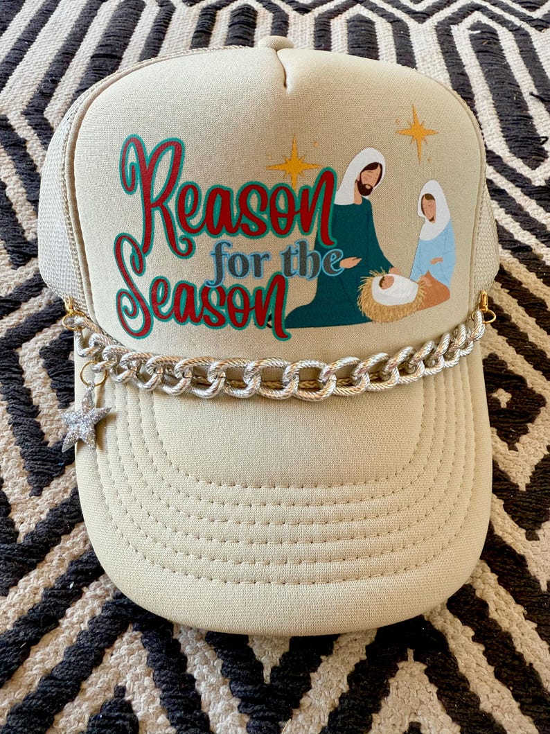 May include: A beige trucker hat with a gold chain trim and a star charm. The front of the hat features a Christmas nativity scene with the text "Reason for the Season".