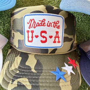 May include: Camouflage trucker hat with a mesh back and a patch that reads "Made in the U.S.A." in red script. The patch is outlined in blue. Three star-shaped patches in red, white, and blue are on the brim.