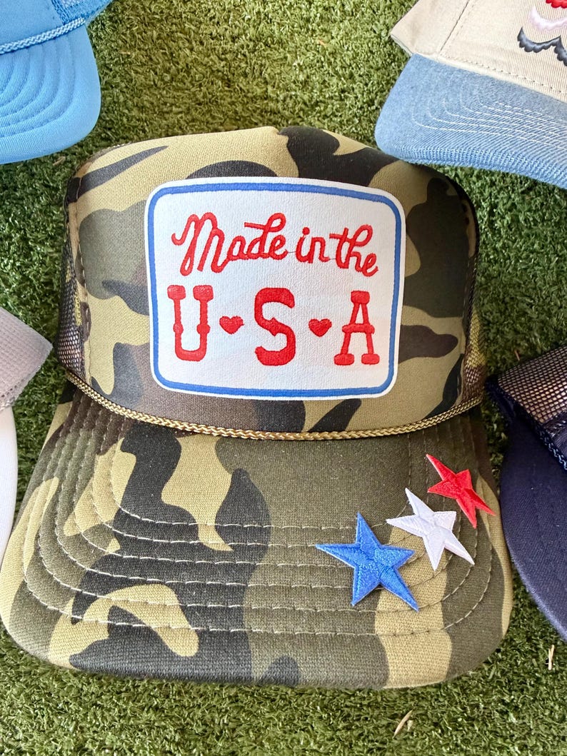 May include: Camouflage trucker hat with a mesh back and a patch that reads "Made in the U.S.A." in red script. The patch is outlined in blue. Three star-shaped patches in red, white, and blue are on the brim.