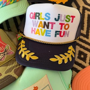 May include: A collection of trucker hats with various designs. The primary hat is navy blue with a white front panel featuring the text "GIRLS JUST WANT TO HAVE FUN" in colorful letters. It has gold leaf accents and a gold chain.