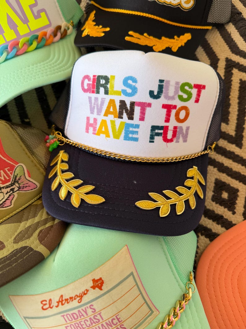 May include: A collection of trucker hats with various designs. The primary hat is navy blue with a white front panel featuring the text "GIRLS JUST WANT TO HAVE FUN" in colorful letters. It has gold leaf accents and a gold chain.
