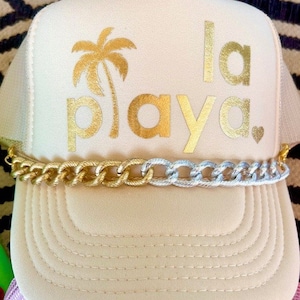 May include: A beige trucker hat with a gold chain trim. The hat has a gold palm tree and the text "la playa" in gold lettering.