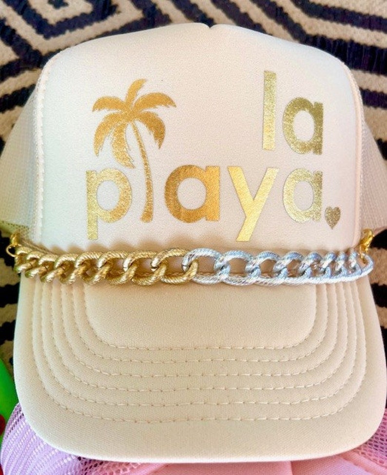 May include: A beige trucker hat with a gold chain trim. The hat has a gold palm tree and the text "la playa" in gold lettering.
