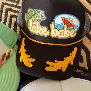 May include: Black trucker hat with a mesh back and a curved brim. The front features a graphic of a green fish, a boat, and the text "lake babe" in yellow script. The hat has yellow embroidered accents.