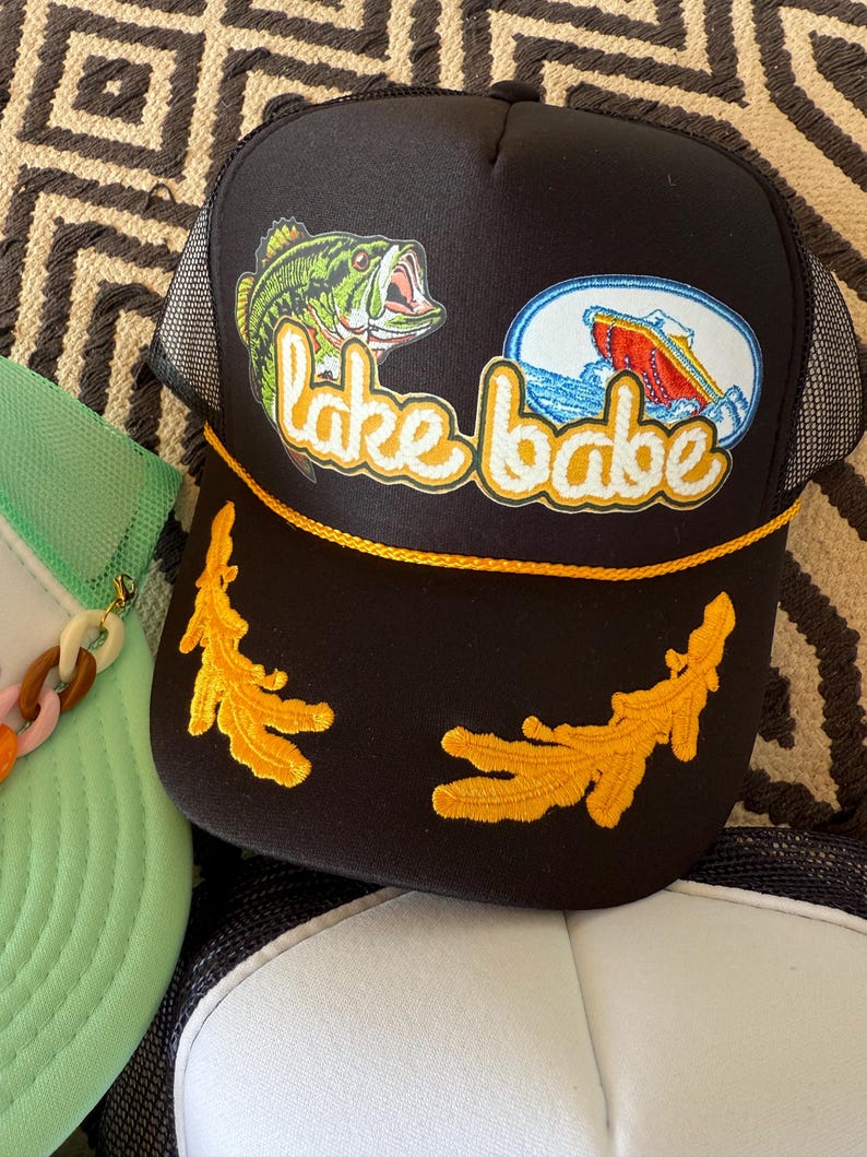 May include: Black trucker hat with a mesh back and a curved brim. The front features a graphic of a green fish, a boat, and the text "lake babe" in yellow script. The hat has yellow embroidered accents.