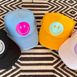 May include: Four trucker hats with different colored mesh and smiley face designs. The hats are blue, yellow, black, and white with pink, light blue, white, and purple smiley faces.